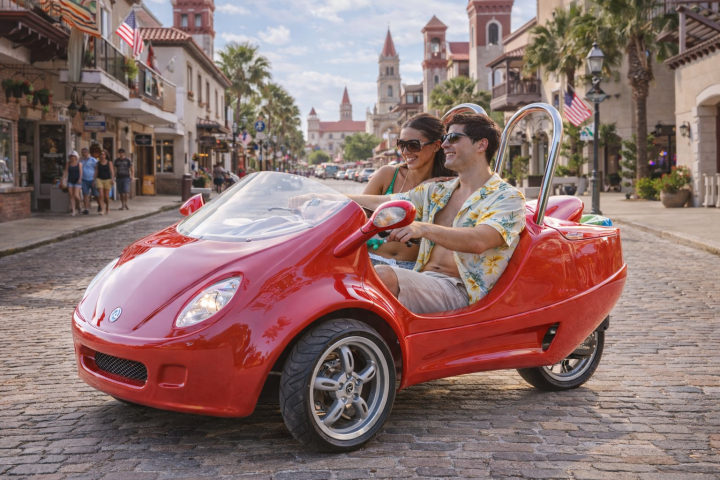 Couple driving a red three-wheeled vehicle on a cobblestone street.