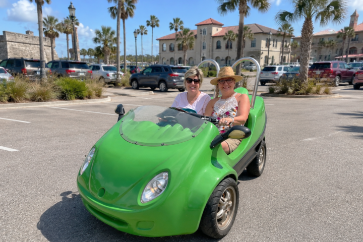 Two people in a small green car in a sunny parking lot with palm trees.