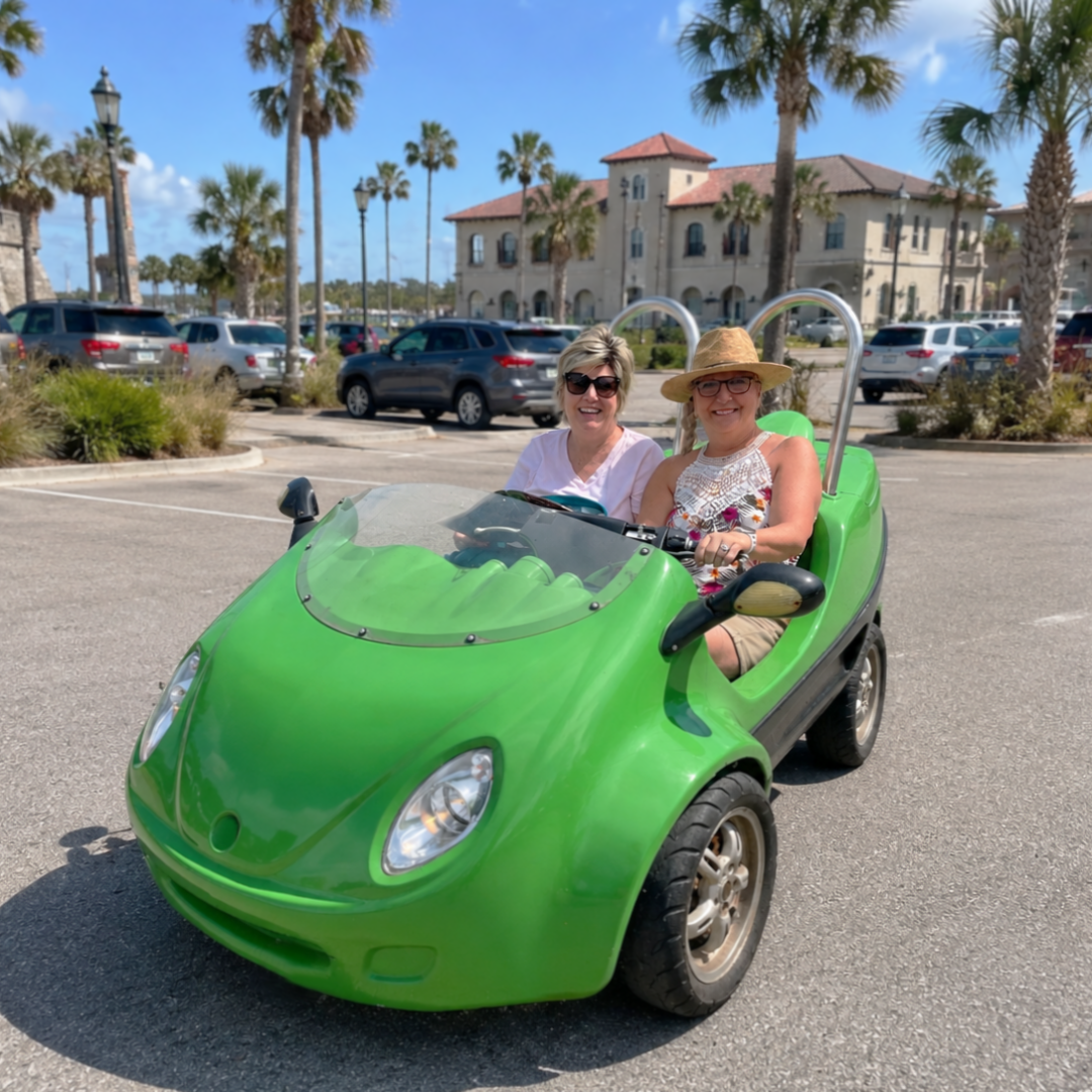 Two people in a small green car in a sunny parking lot with palm trees.