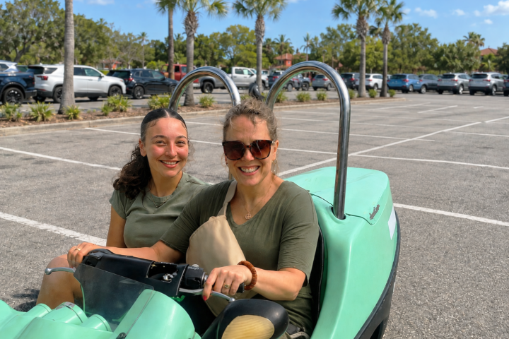 Two women smiling in a green go-kart in a parking lot with palm trees.