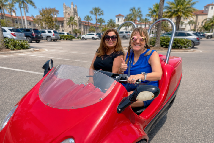 Two women smiling in a red open-air car in a sunny parking lot.