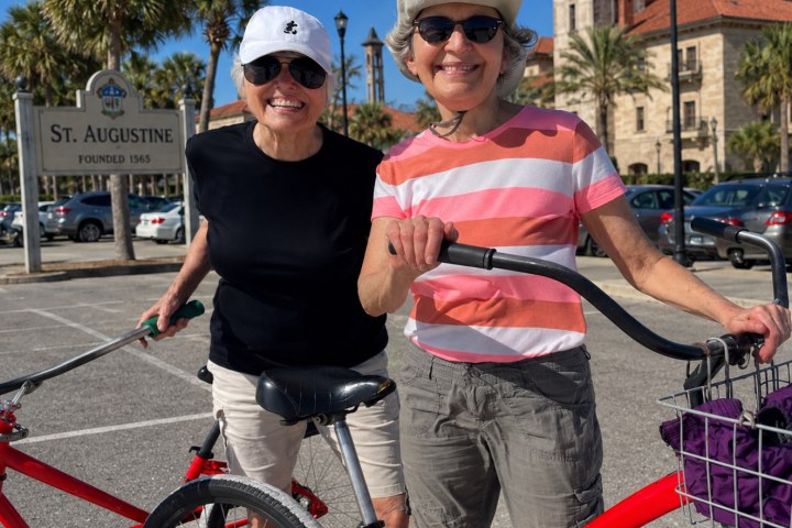 Two people smiling with red bikes in a sunny parking area near a St. Augustine sign.