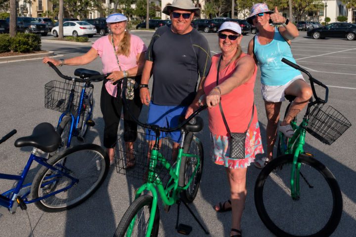 Four people with bicycles in a sunny parking lot, wearing hats and casual clothes.