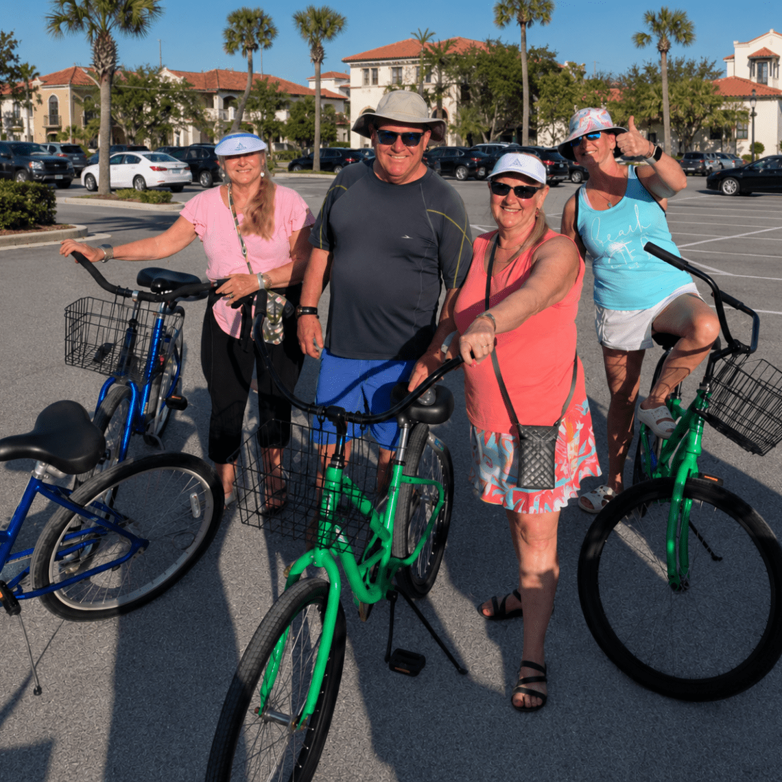 Four people with bicycles in a sunny parking lot, wearing hats and casual clothes.