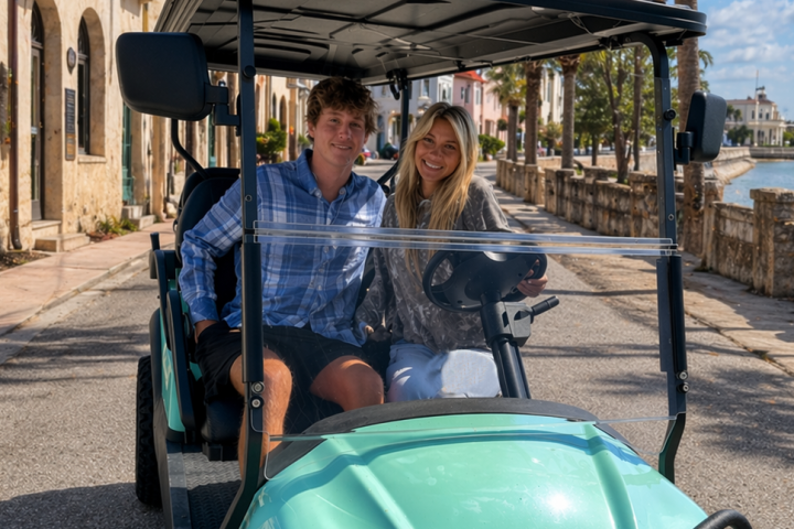 Two people smiling in a turquoise golf cart on a sunny street.