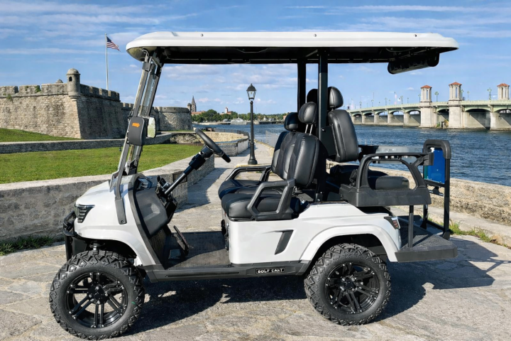 White golf cart parked by a waterfront with bridge and flag in background.