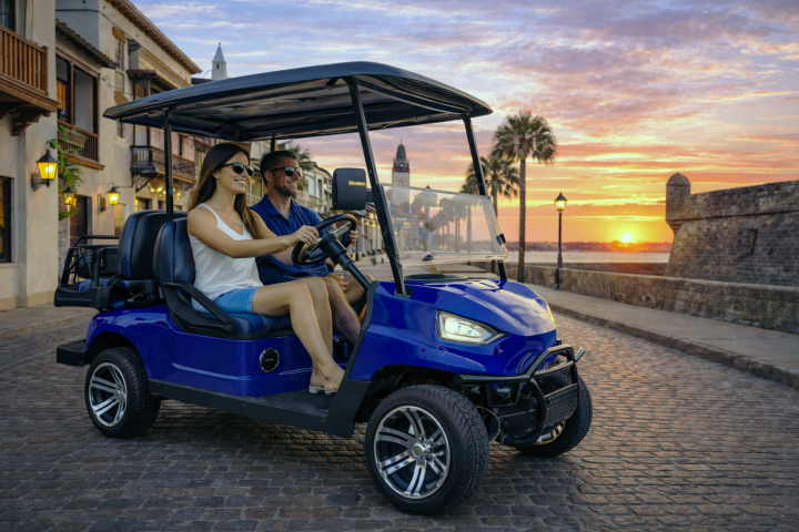 Couple driving blue golf cart on cobblestone street at sunset.