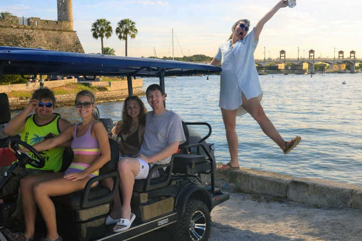 Four people in a golf cart near water, a woman stands joyfully on a wall holding a camera.