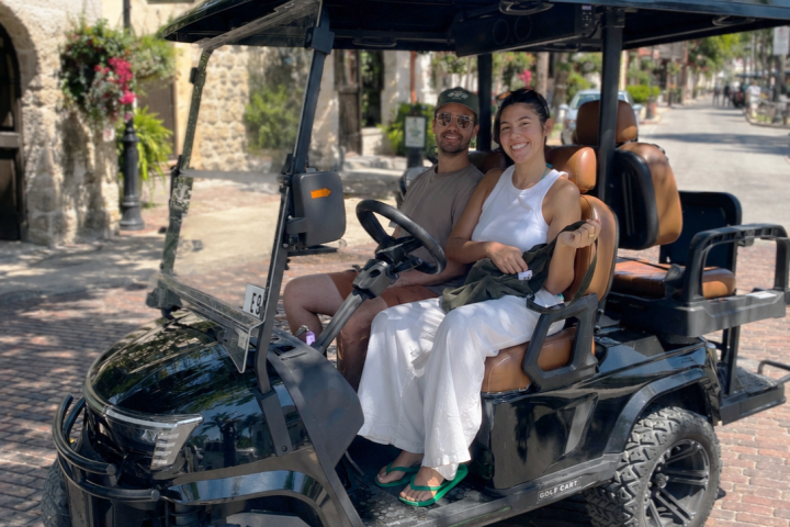 Two smiling people in a black golf cart on a sunny cobblestone street.