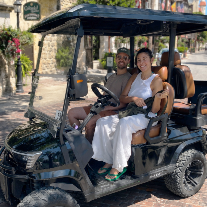 Two smiling people in a black golf cart on a sunny cobblestone street.