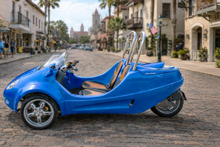 Blue three-wheeled vehicle on cobblestone street with historic buildings.