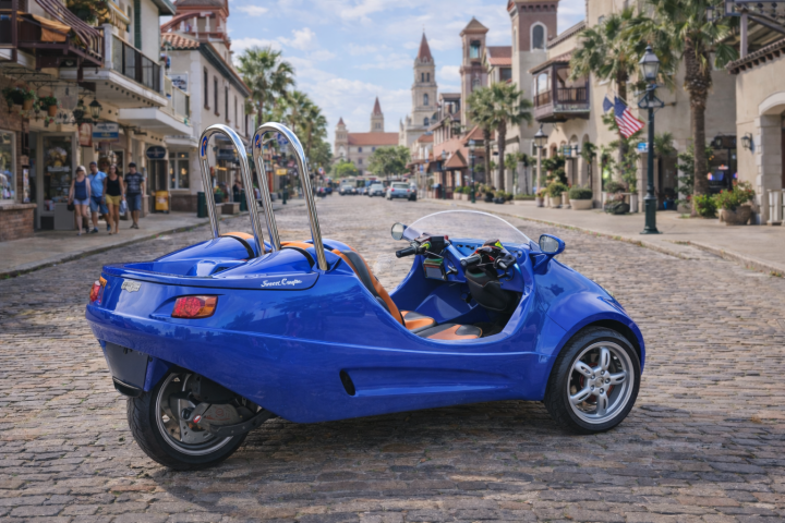Blue three-wheeled vehicle parked on cobblestone street in a scenic town setting.