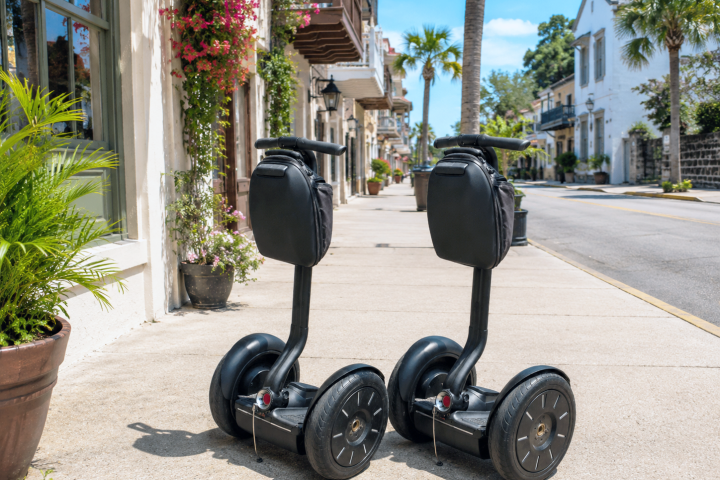 Two Segways parked on a sidewalk in a sunny street with palm trees and buildings.