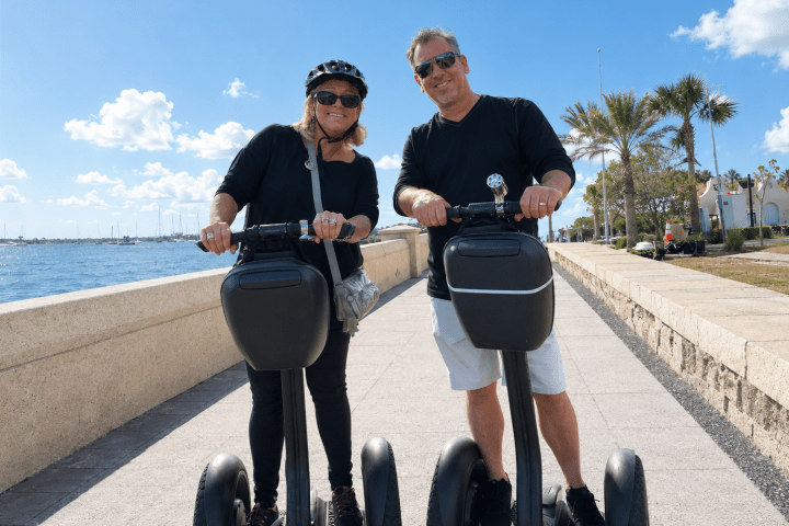 Two people on Segways by a waterfront path, wearing casual clothes and sunglasses.