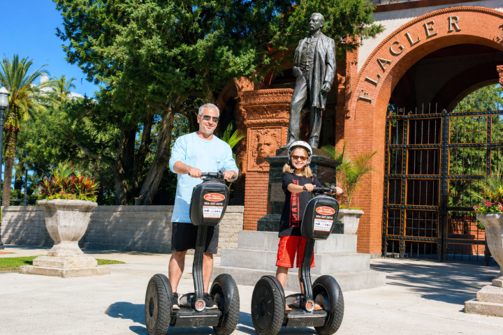 Two people on Segways in front of a statue outside a gate.
