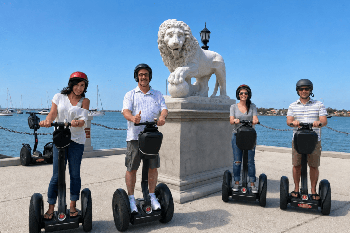 Four people on Segways pose by a lion statue near water on a sunny day.