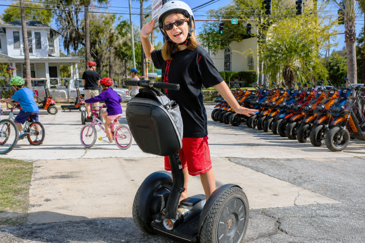 Child on a Segway waving, wearing a helmet, with bikes and trees in the background.
