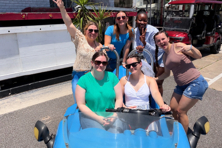 Six smiling people pose around a blue cart, outdoors on a sunny day.
