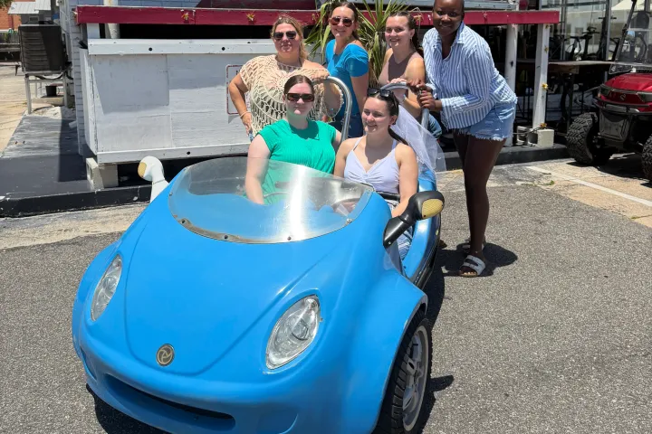Group of six people posing with a small blue car in a sunny outdoor setting.