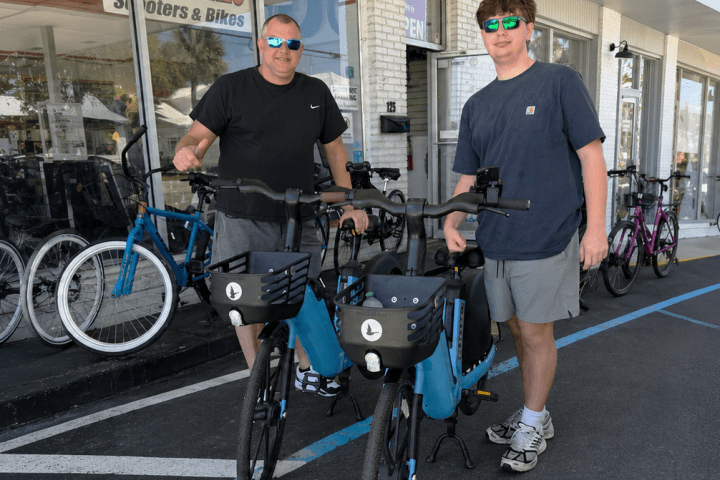 Two people with e-bikes outside a store on a sunny day.