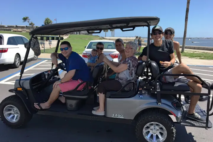 Group of people smiling in a parked golf cart near a waterfront.