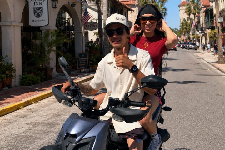 Two people on a scooter, smiling on a sunny street lined with palm trees.