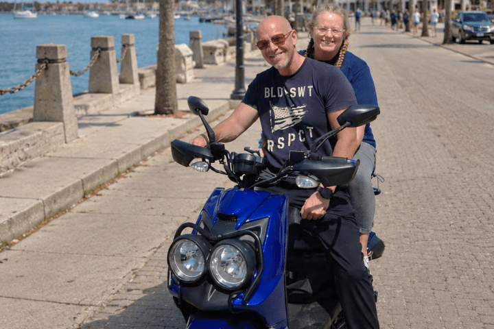 Two people smiling on a blue scooter near a waterfront pathway.