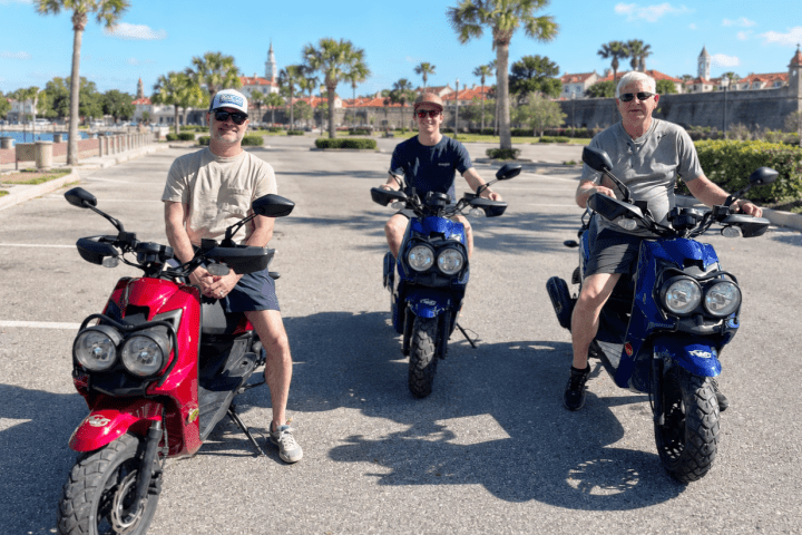 Three people on motor scooters in a parking lot with trees in the background.