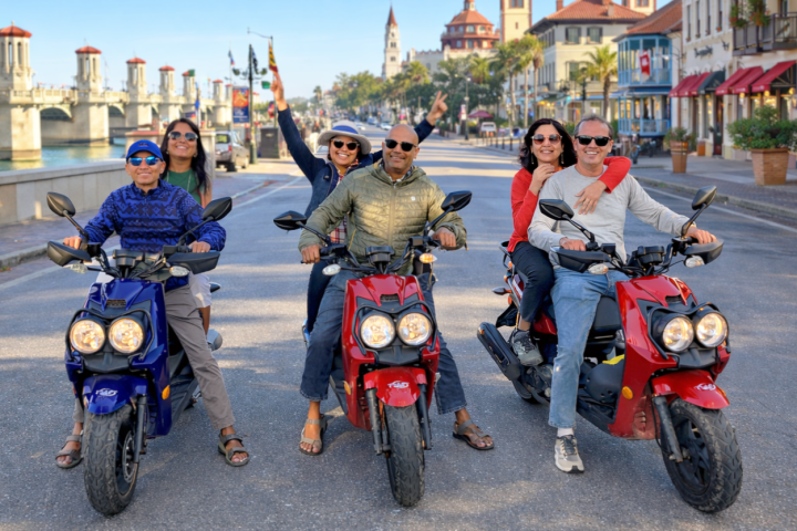 Group of six people on scooters posing on a sunny street.
