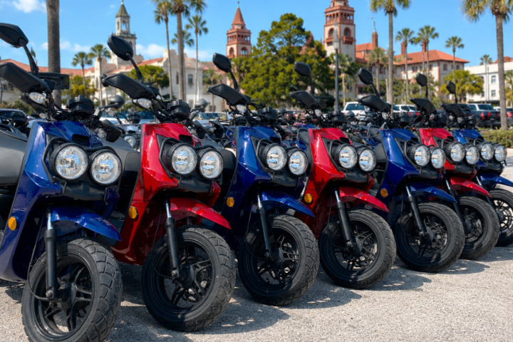 Row of blue and red scooters parked on a sunny street with palm trees and historic buildings.