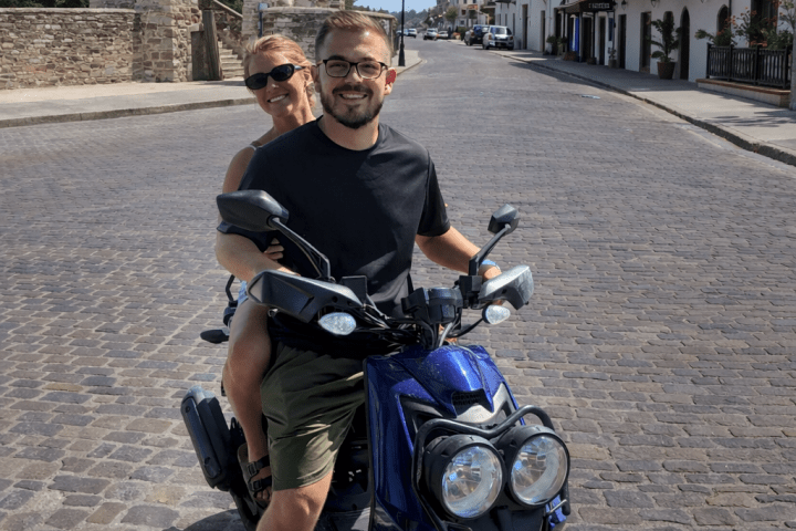 Two people smiling on a blue scooter in a cobblestone street.