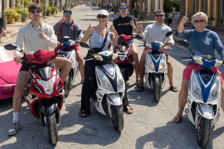 Five people on scooters giving thumbs up on a sunny street.
