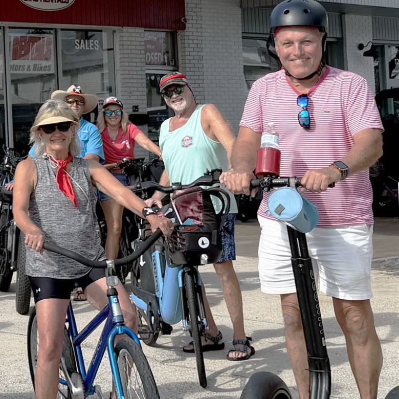 Group of people with bikes and scooters outside a rental shop.